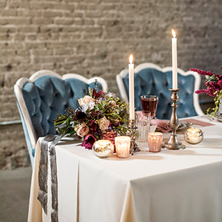 A tablescape filled with winter wedding colors: two plush blue velvet chairs at a table with taper candles in metallic holders, mercury glass votives, and burgundy flower arrangements with greenery and grey ribbon