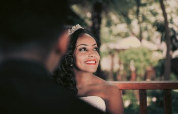 A woman with long wavy black hair wearing a tiara and wedding dress looks up, smiling