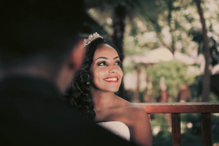 A woman with long wavy black hair wearing a tiara and wedding dress looks up, smiling