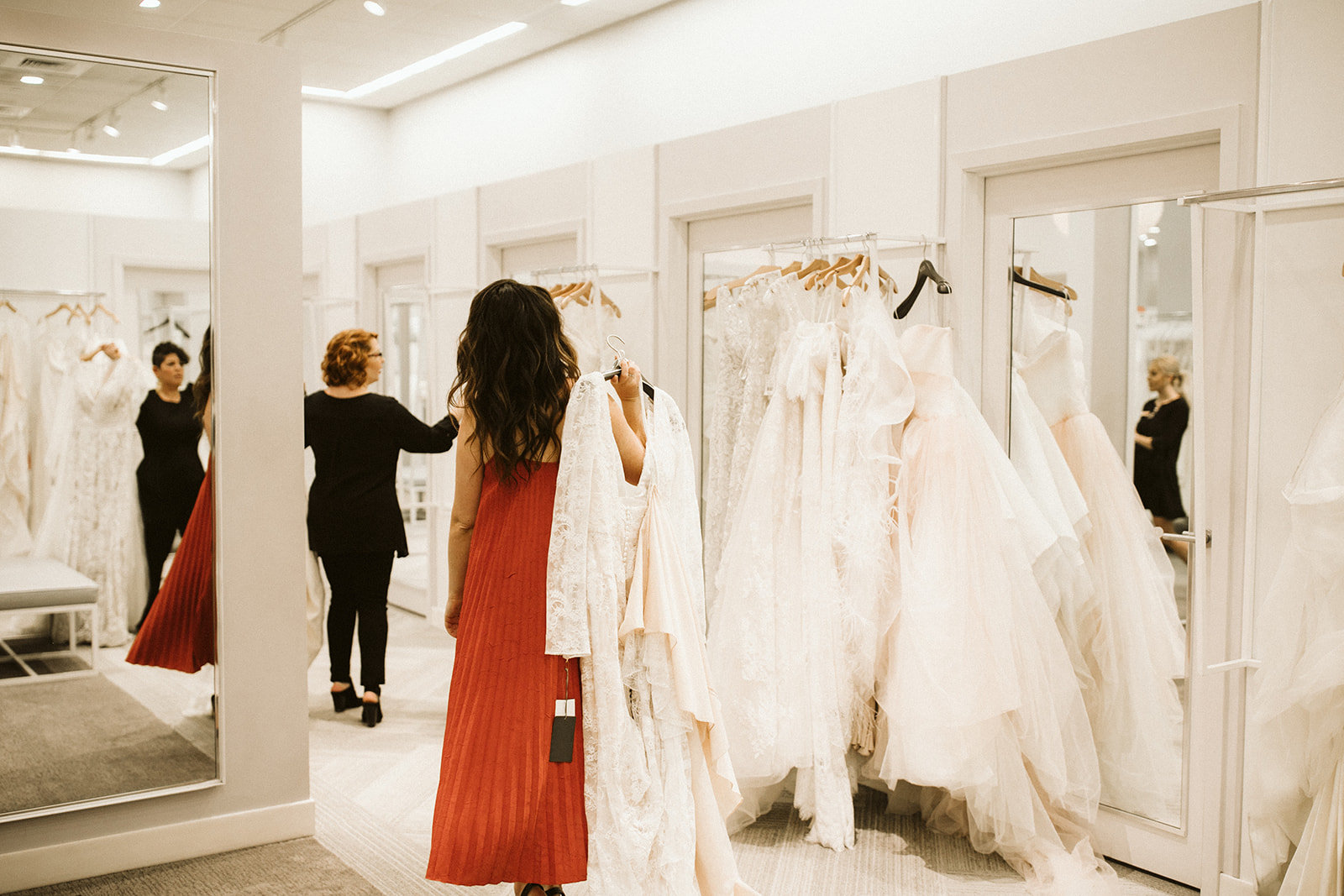 A brunette woman in an orange dress carries a wedding gown toward a dressing room at David's Bridal