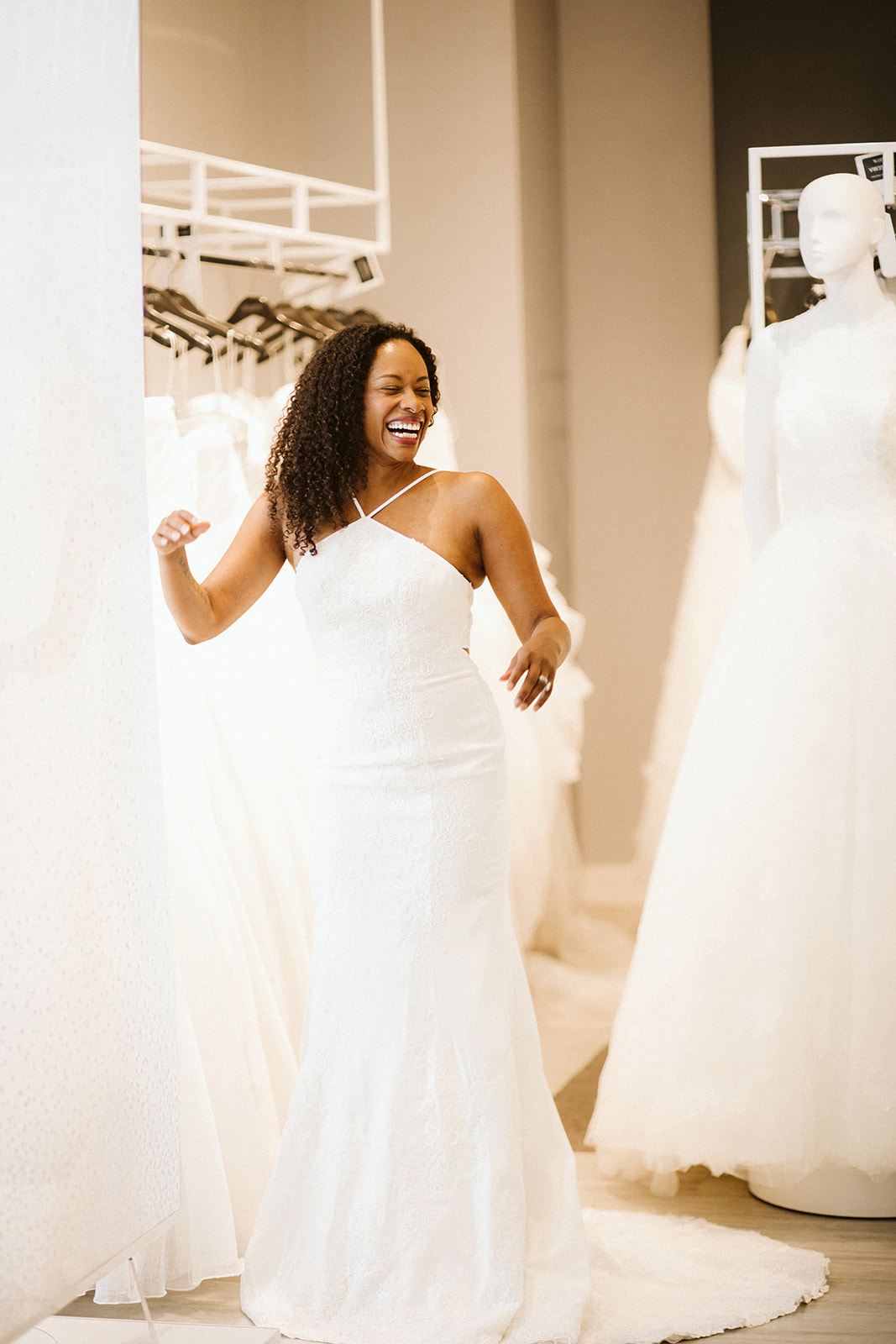 A woman with coiled dark laughs near a mannequin wearing a wedding gown in the showroom at David's Bridal while she wears a halter-neck fitted white wedding dress with a small train