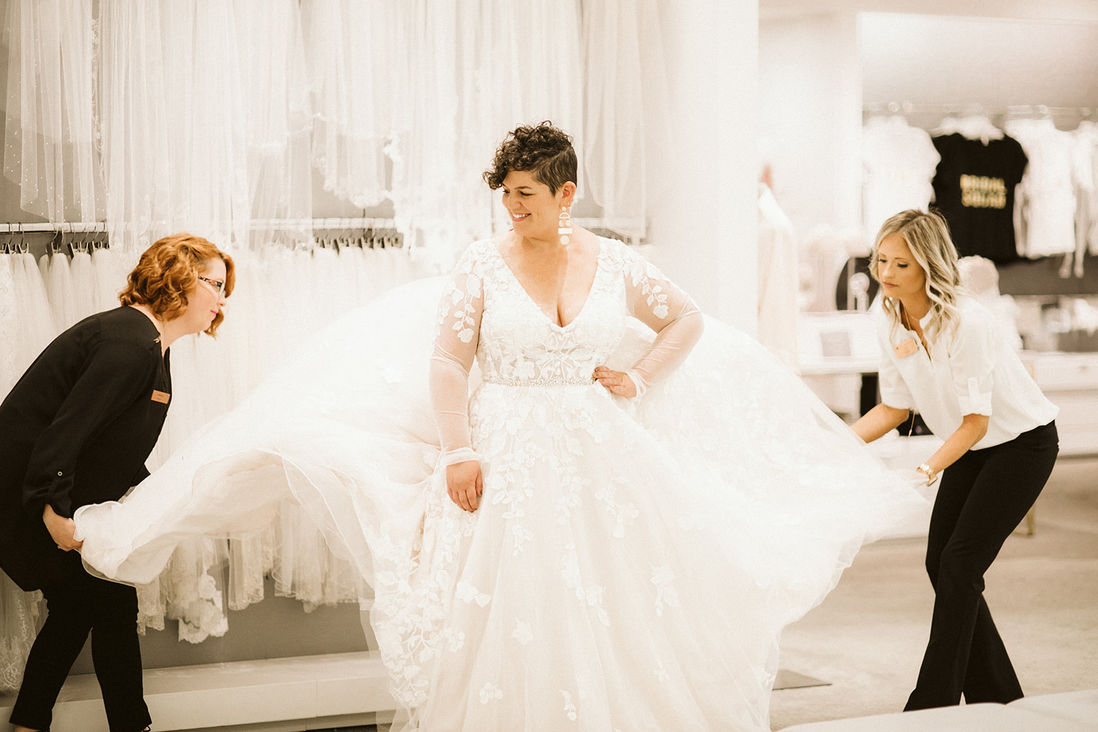 Two sales assistants fluff the skirt of a long sleeve wedding dress worn by a short-haired brunette woman at David's Bridal