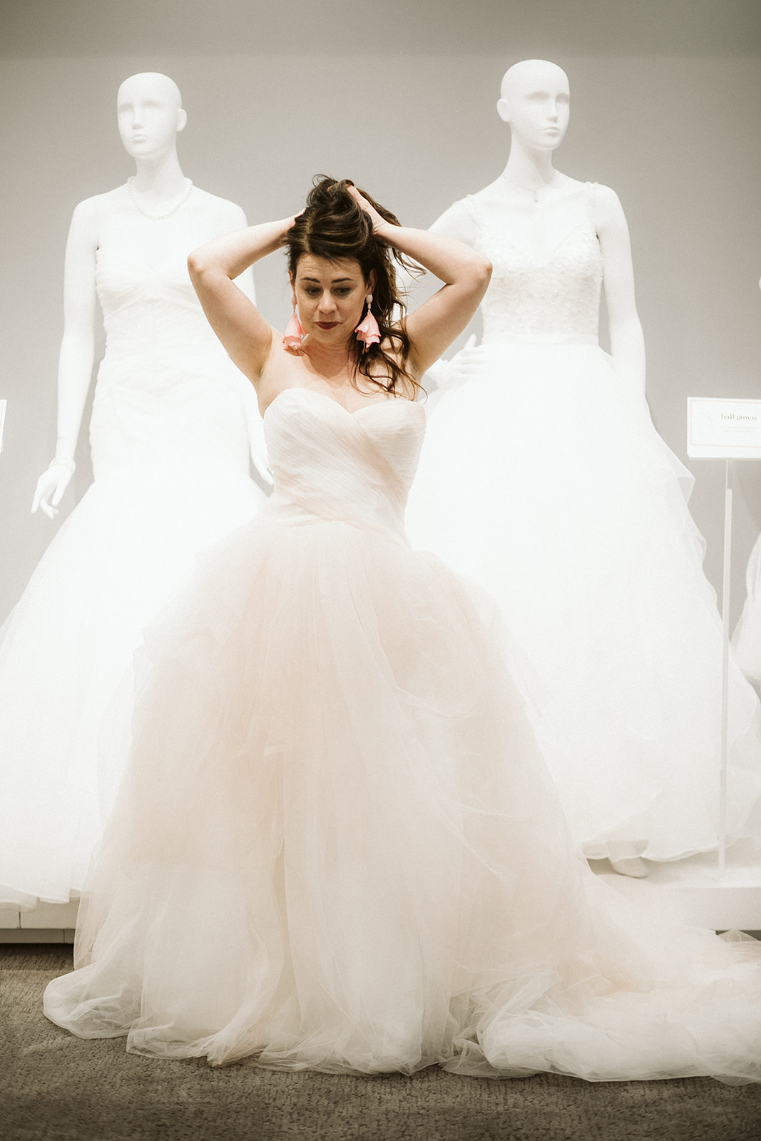 A a brunette woman in a strapless tulle ball gown holds her hair above her head as she stands in front of two mannequins in wedding dresses at David's Bridal