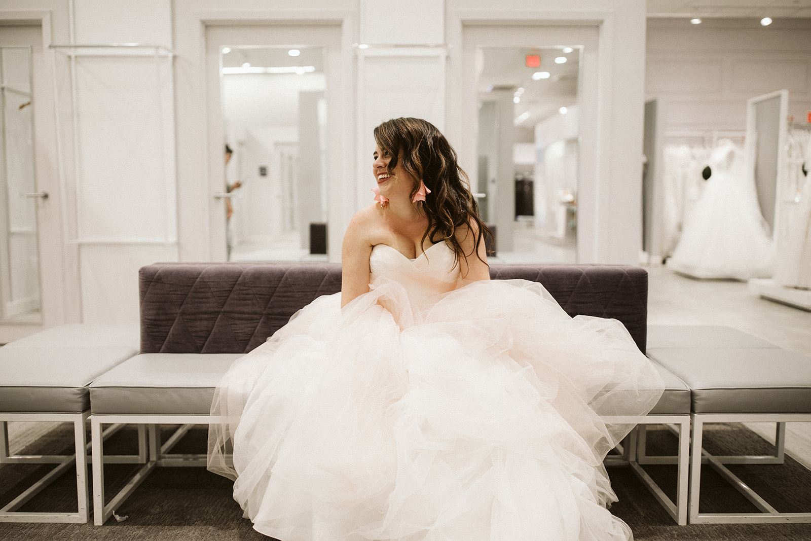 A woman with long dark hair sits in a blush tulle ball gown on a modern grey sofa in the brightly lit fitting room at David's Bridal