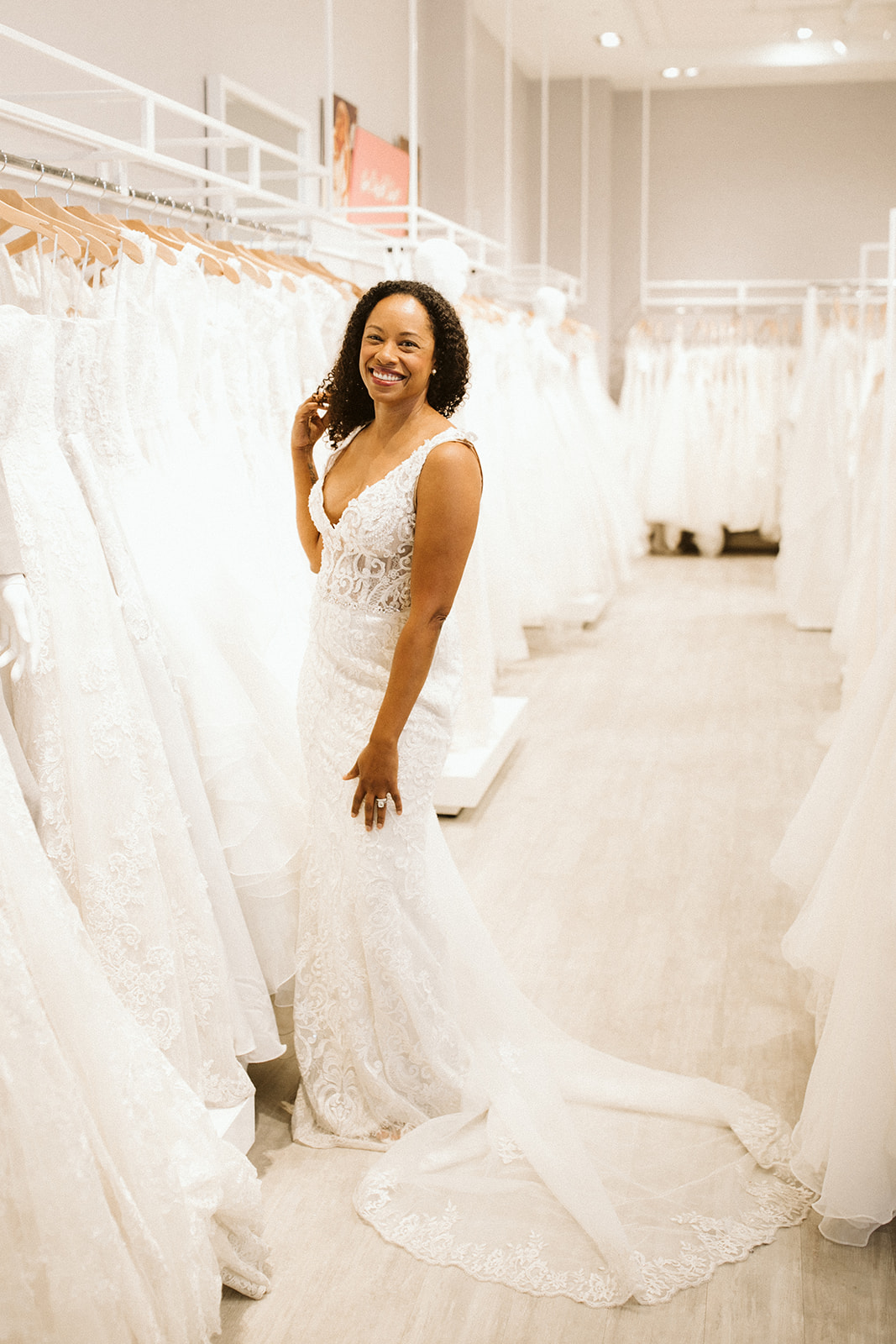 A woman with coiled dark hair wears a lace sleeveless wedding gown with a train and smiles as she touches her hair in the show room at David's Bridal