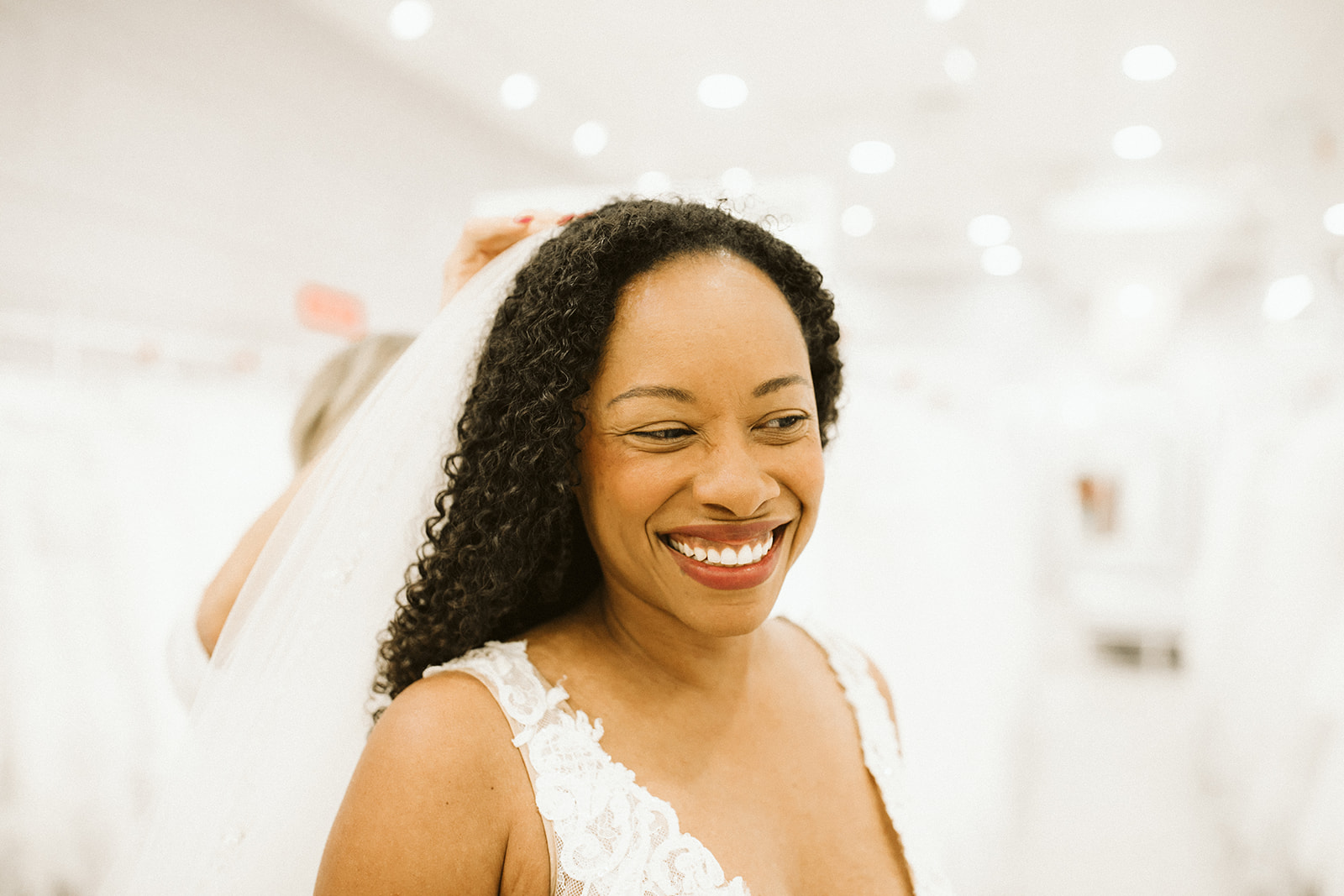 A woman with coiled dark hair smiles as she has a veil pinned to her head at David's Bridal