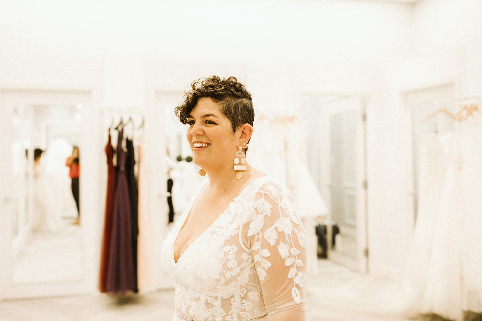 A woman with short brown hair smiles as she tries on a lace wedding dress at David's Bridal