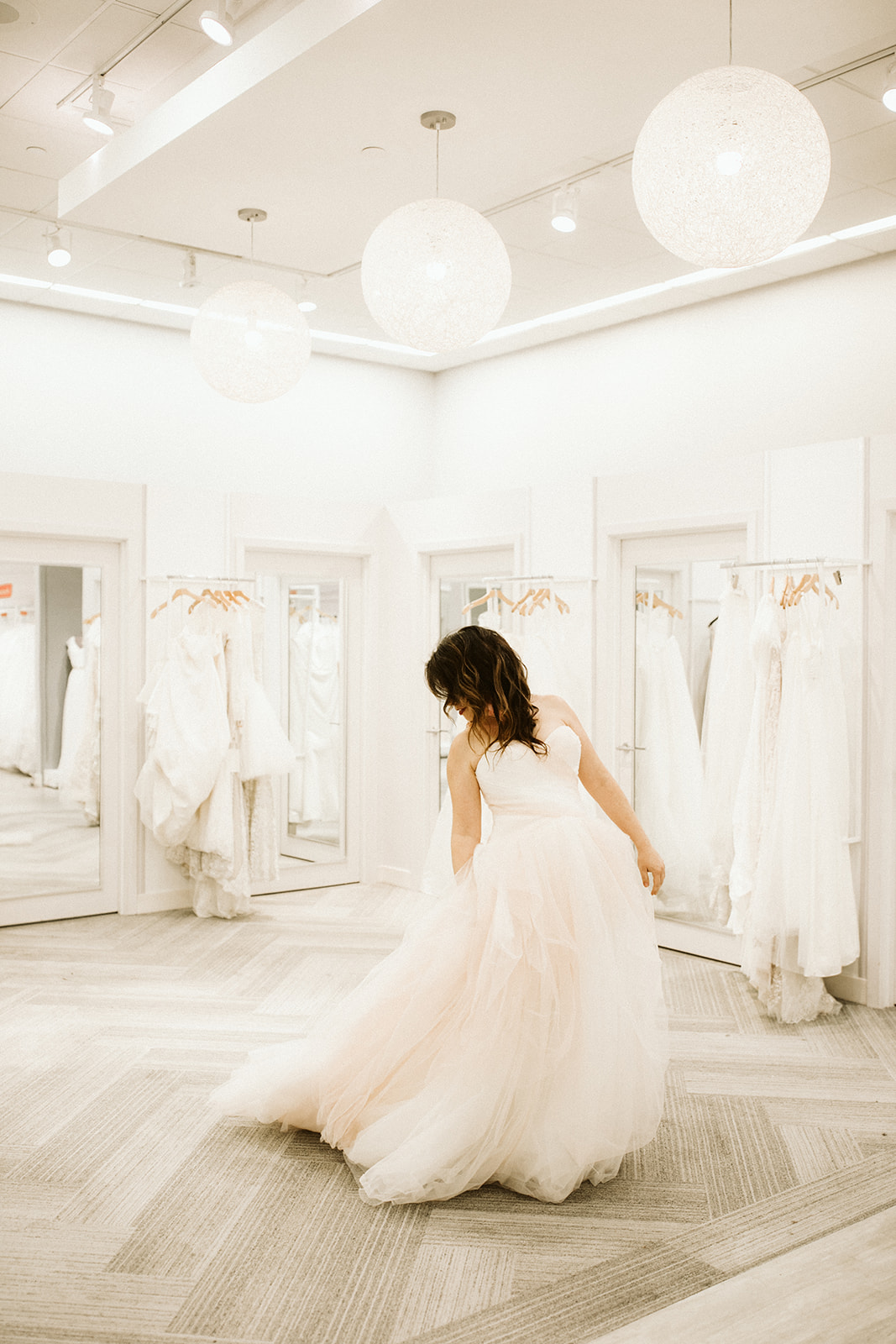 A woman in a blush tulle ball gown looks back and adjusts her skirt in a brightly lit modern fitting room at David's Bridal