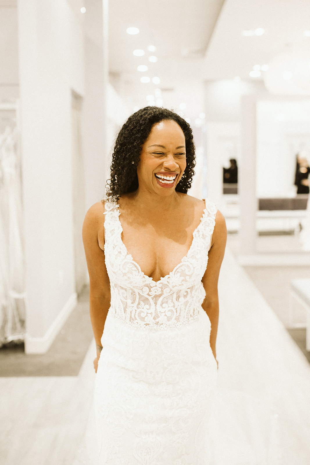 A woman with coiled dark hair laughs in the showroom at David's Bridal as she wears a sleeveless lace wedding dress with a scoop plunge-v-neckline