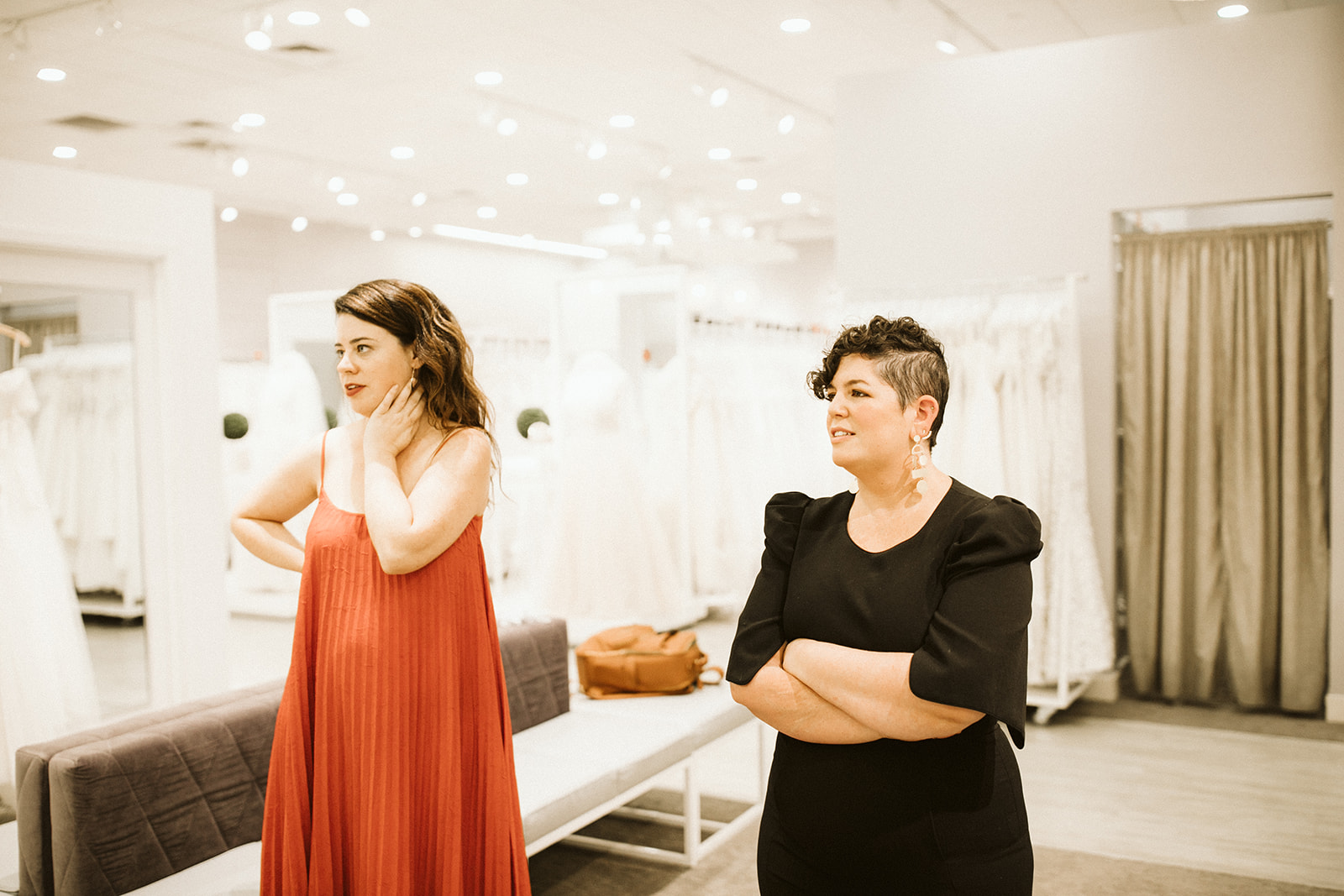 Two brunette women, one with long hair wearing an orange dress, the other with short hair, wearing a black dress, stand in a modern bridal showroom at David's Bridal
