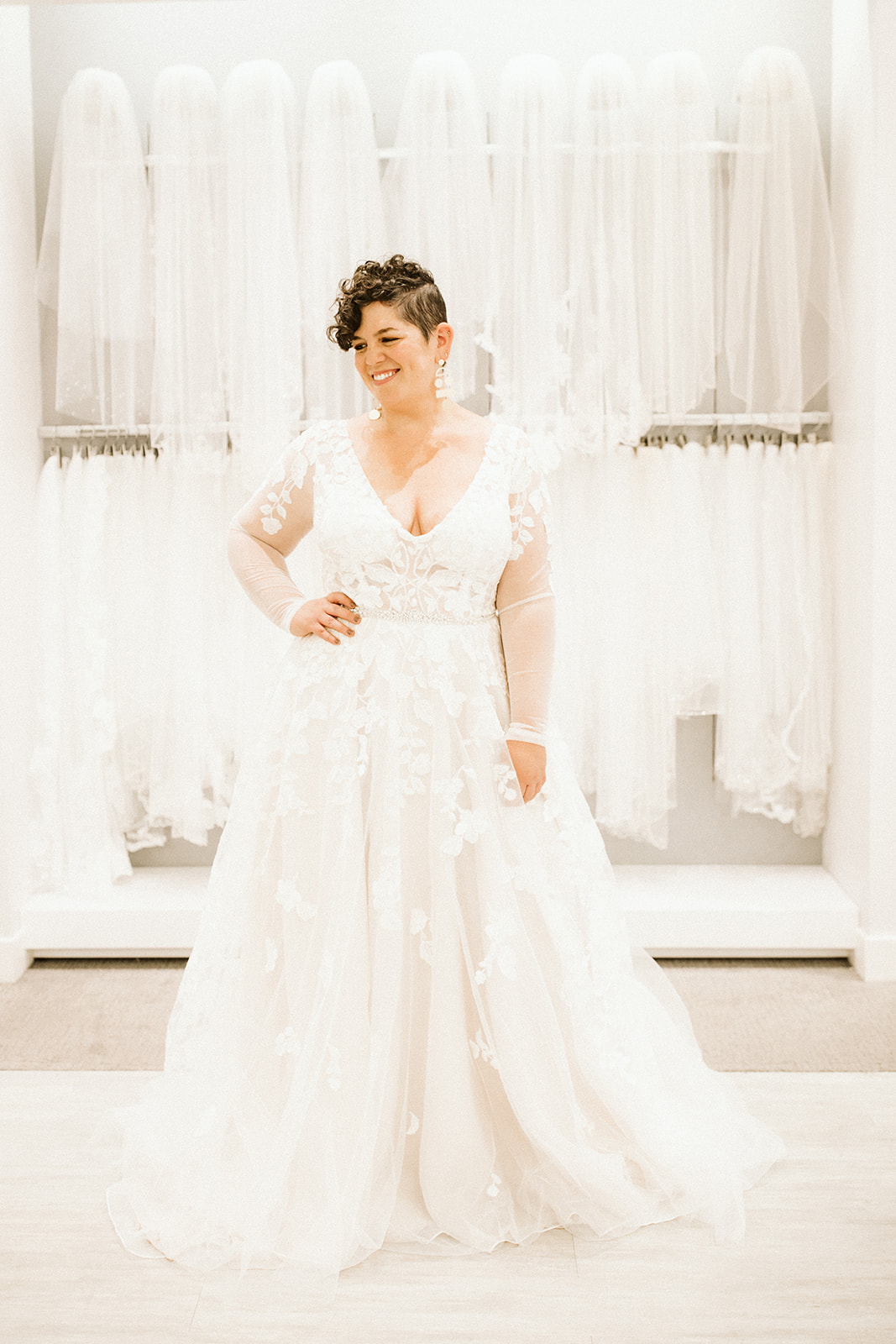 A woman with short brown hair stands in a long sleeve lace wedding gown in front of a wall of veils at David's Bridal