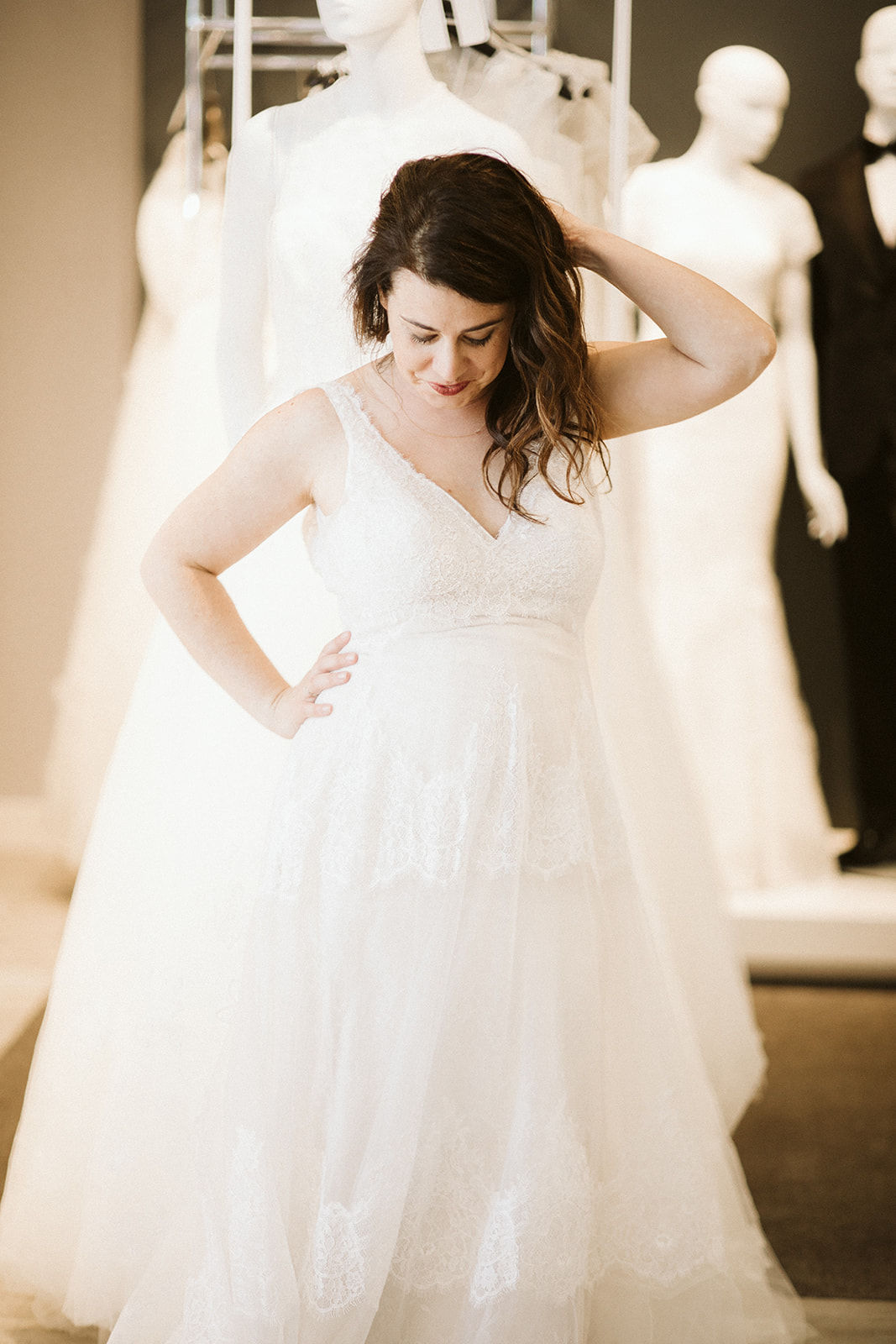 A woman with long brown hair touches her head as she looks down while wearing a sleeveless v-neck wedding gown with lace appliqués at David's Bridal