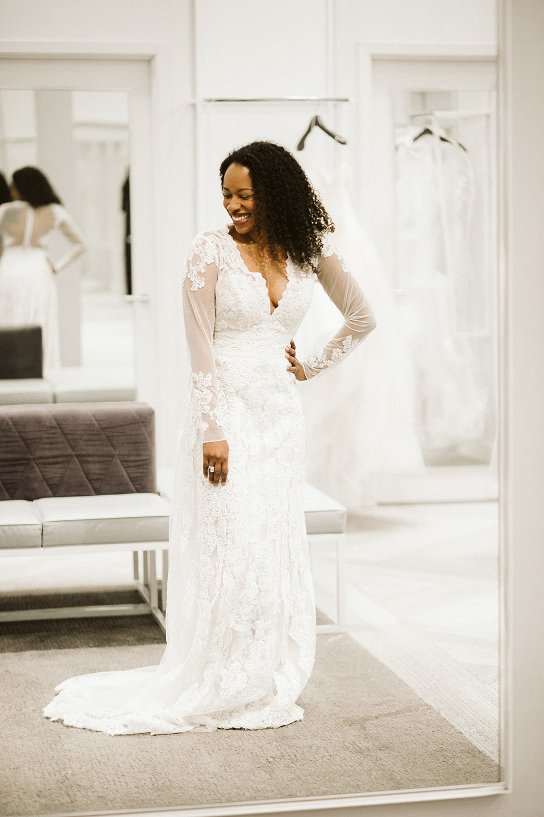 A woman with coiled dark hair smiles with her hand on her hip as she stands in front of a mirror wearing a long sleeve deep-v neck wedding gown in the modern fitting room at David's Bridal
