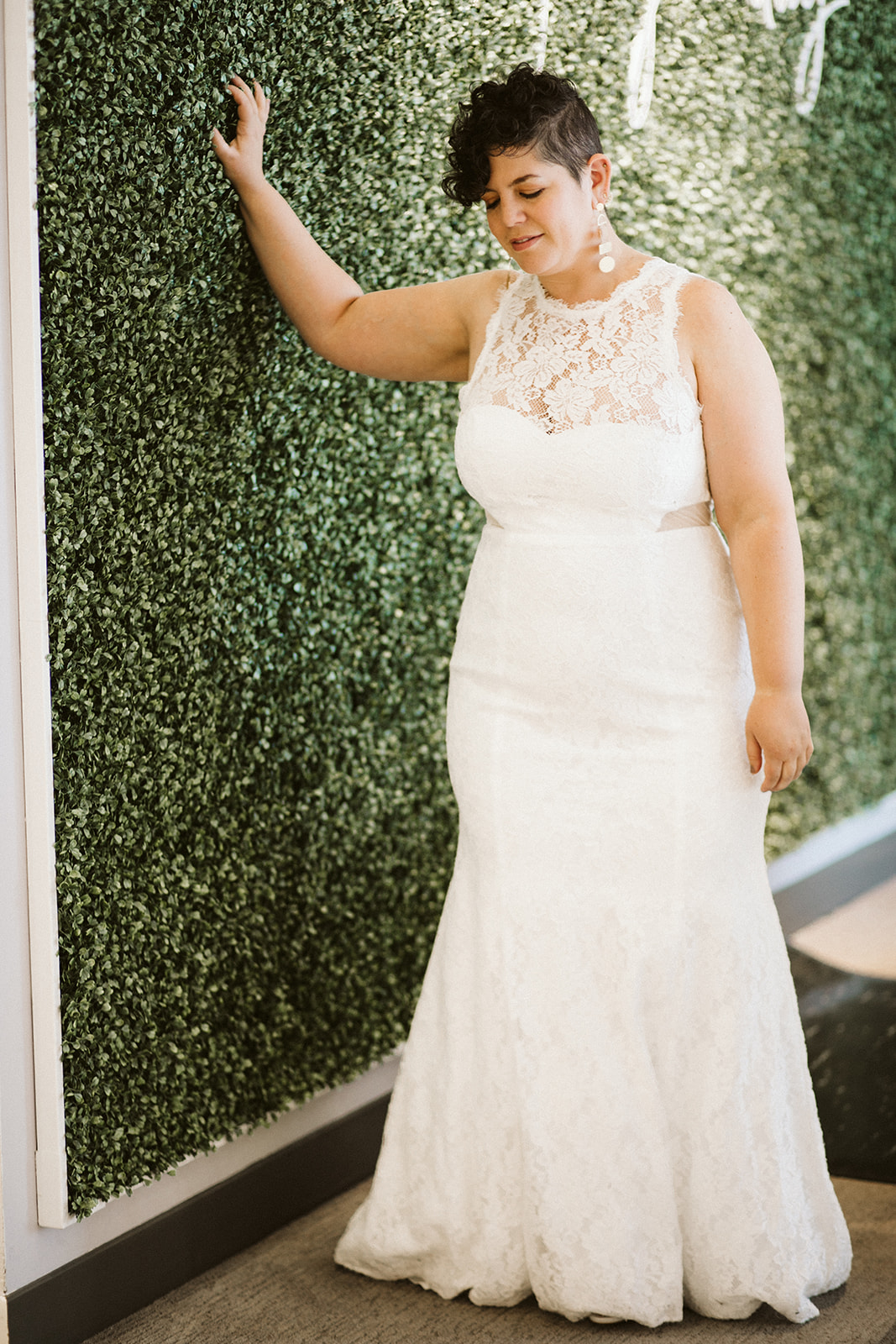 A curvy brunette woman with short hair stands in front of a greenery wall in a sleeveless lace wedding gown with an illusion neckline and cutouts at the side waist at David's Bridal