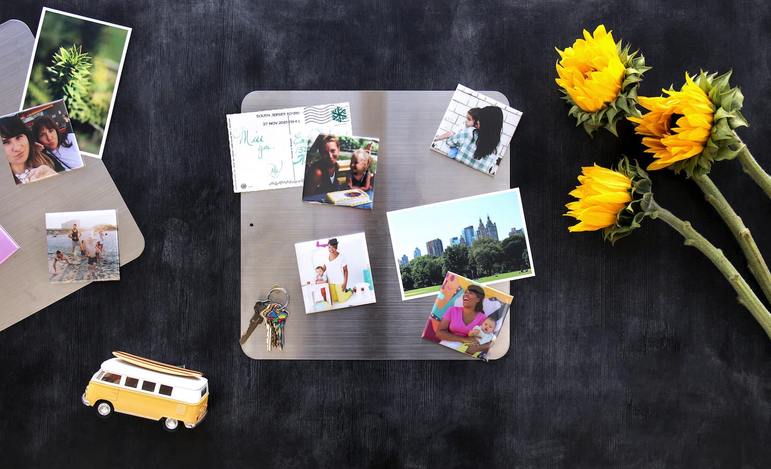 a silver magnet board covered in postcards, photos and photo magnets on a black background with sunflowers next to it