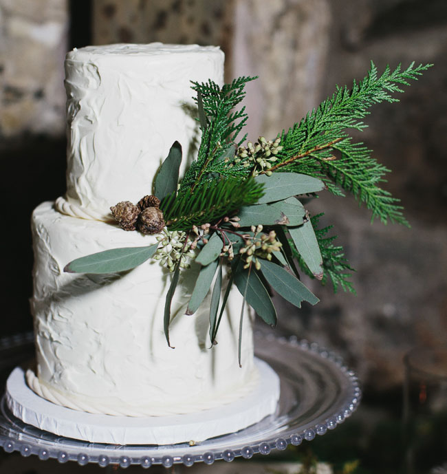 frosted tiered cake in winter wedding colors sitting on table: white cake with green foliage decorations and pinecones