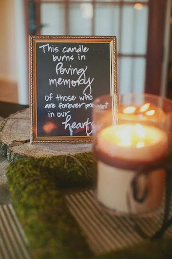 wedding ideas to celebrate those who have passed—a memorial sign that reads "this candle burns in loving memory of those who are forever present in our hearts" and a lit pilar candle in a glass holder