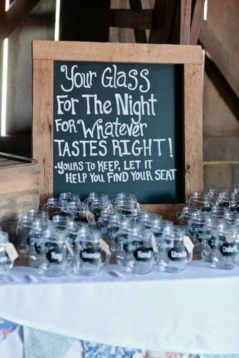 wedding ideas for cups—customizable mason jars. A sign reads "Your Glass for the night for whatever tastes right! Yours to keep, let it help you find your seat" above rows of mason jars with handles with names written on the side