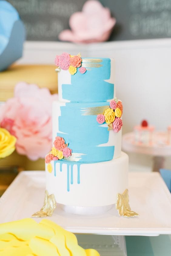 wedding ideas for cakes—a hand-painted cake with blue and silver streaks on three tier cake with gold leaf "feet" and pink, yellow, and orange edible rosettes 