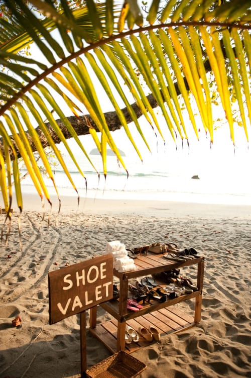 wedding ideas for a shoe valet on the beach—a sign reads "shoe valet" underneath a tropical tree on a sandy beach beside a three tiered shoe rack and small tower of rolled white hand towels