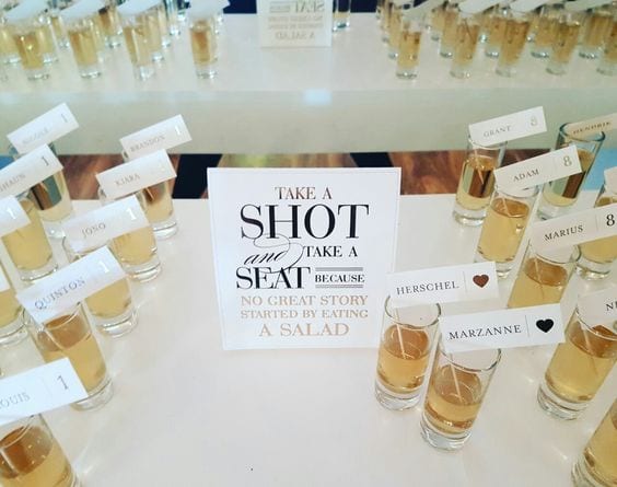 Wedding ideas for a seating chart—shots seating charts with a sign that reads "Take a shot and take a seat because no great story started by eating a salad" with rows of shot glasses with flags with names and table numbers