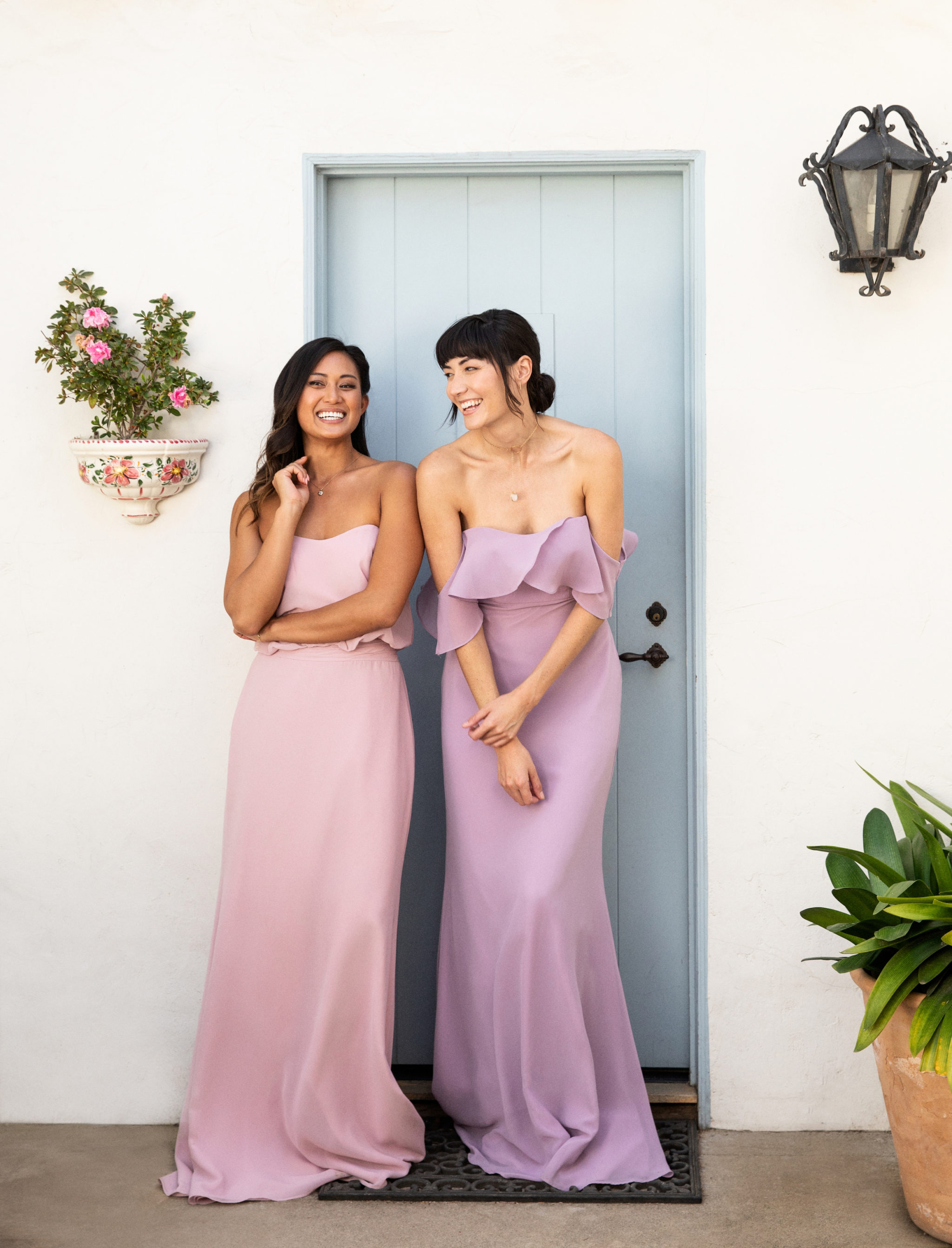 Two women in Brideside bridesmaid dresses laugh in front of a light blue door.