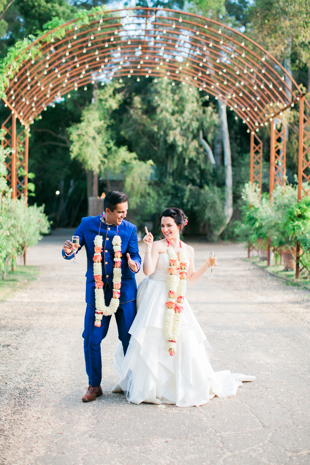 Groom in blue Indian wedding outfit and bride in white dress both wearing long floral garlands dance outdoors under an archway of lights in a photo by Laura Ford