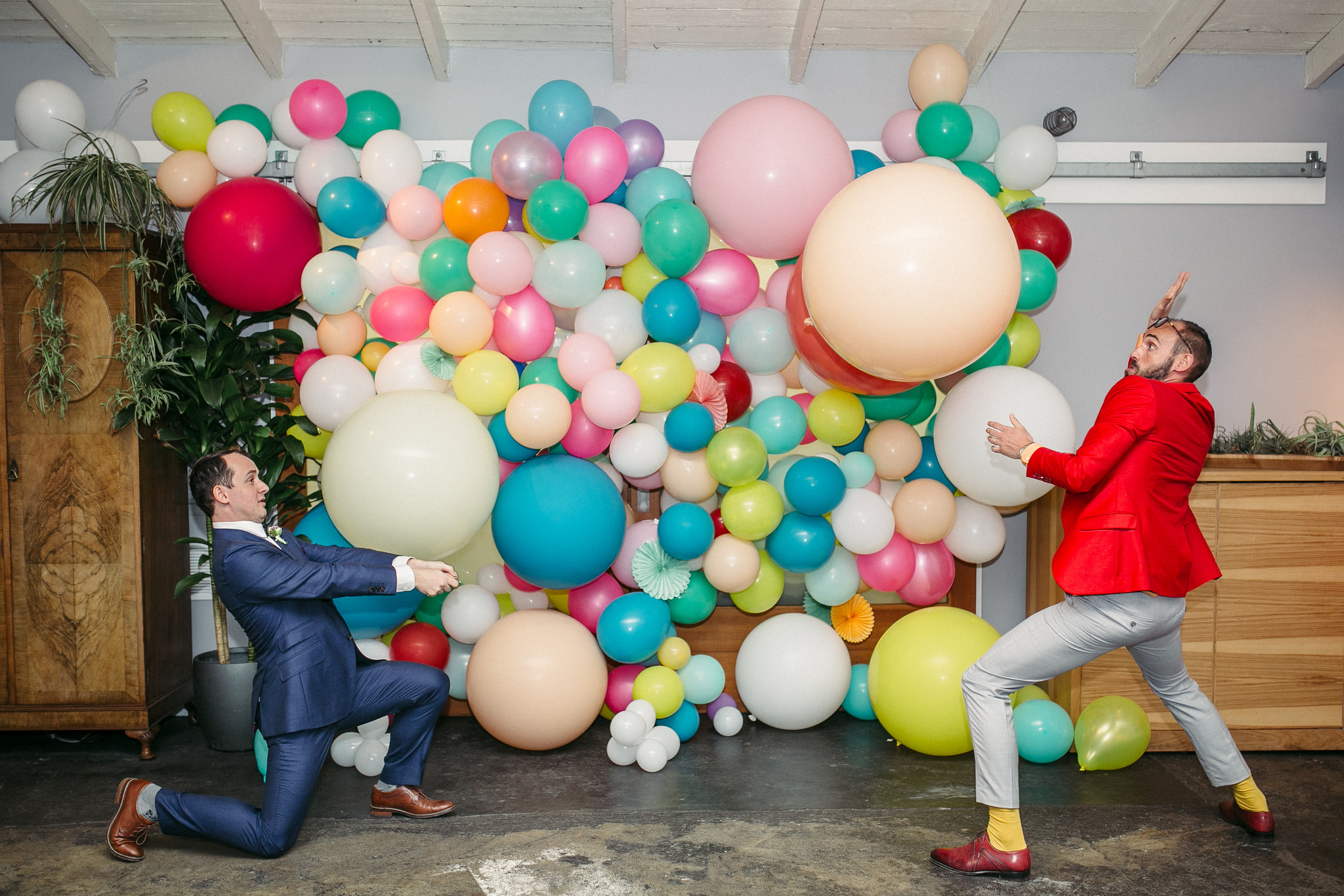 Two men in suits play with giant balloons in front of a colorful balloon installation