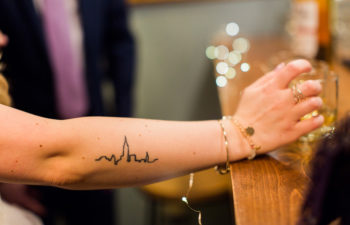 A woman with a line drawing tattoo on her forearm rests her hand on a bar near a whiskey glass. Twinkle lights shine out of focus down the bar.