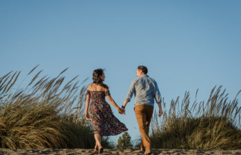 A couple holds hands and walks away from you on a windy sand dune