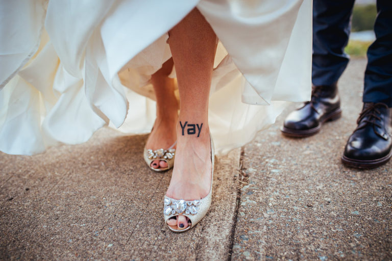 A woman in a wedding dress shows off her ankle tattoo that reads "yay" and her bauble encrusted shoes