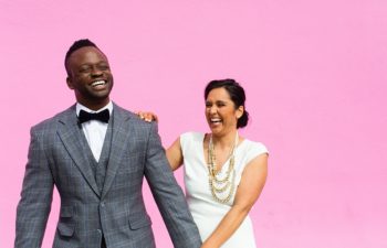 A hetero inter-racial couple holds hands, laughing, standing in their wedding clothes in front of a bright pink wall