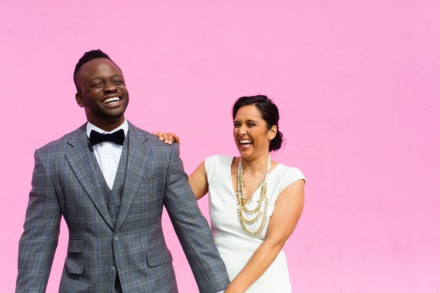 A hetero inter-racial couple holds hands, laughing, standing in their wedding clothes in front of a bright pink wall