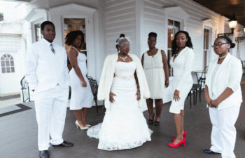 A bride and wedding party, all clad in white, on a veranda in front of a white house