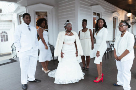 A bride and wedding party, all clad in white, on a veranda in front of a white house