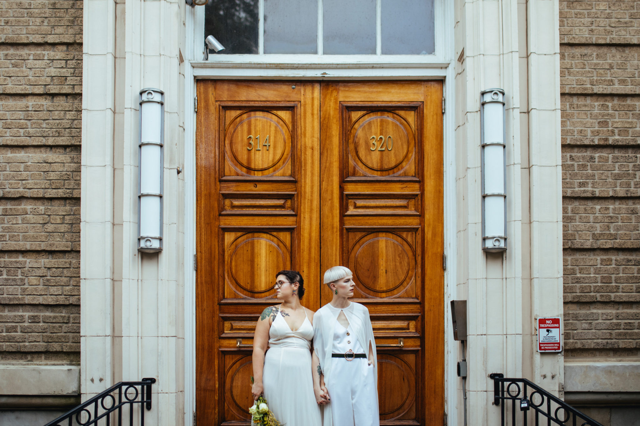 couple stand holding hands in front of big wooden doors on their wedding, one has brown braided hair and tattoos and is wearing a white slip-dress gown while the other has cropped platinum hair and it wearing a white jumpsuit with a chiffon cape over it.