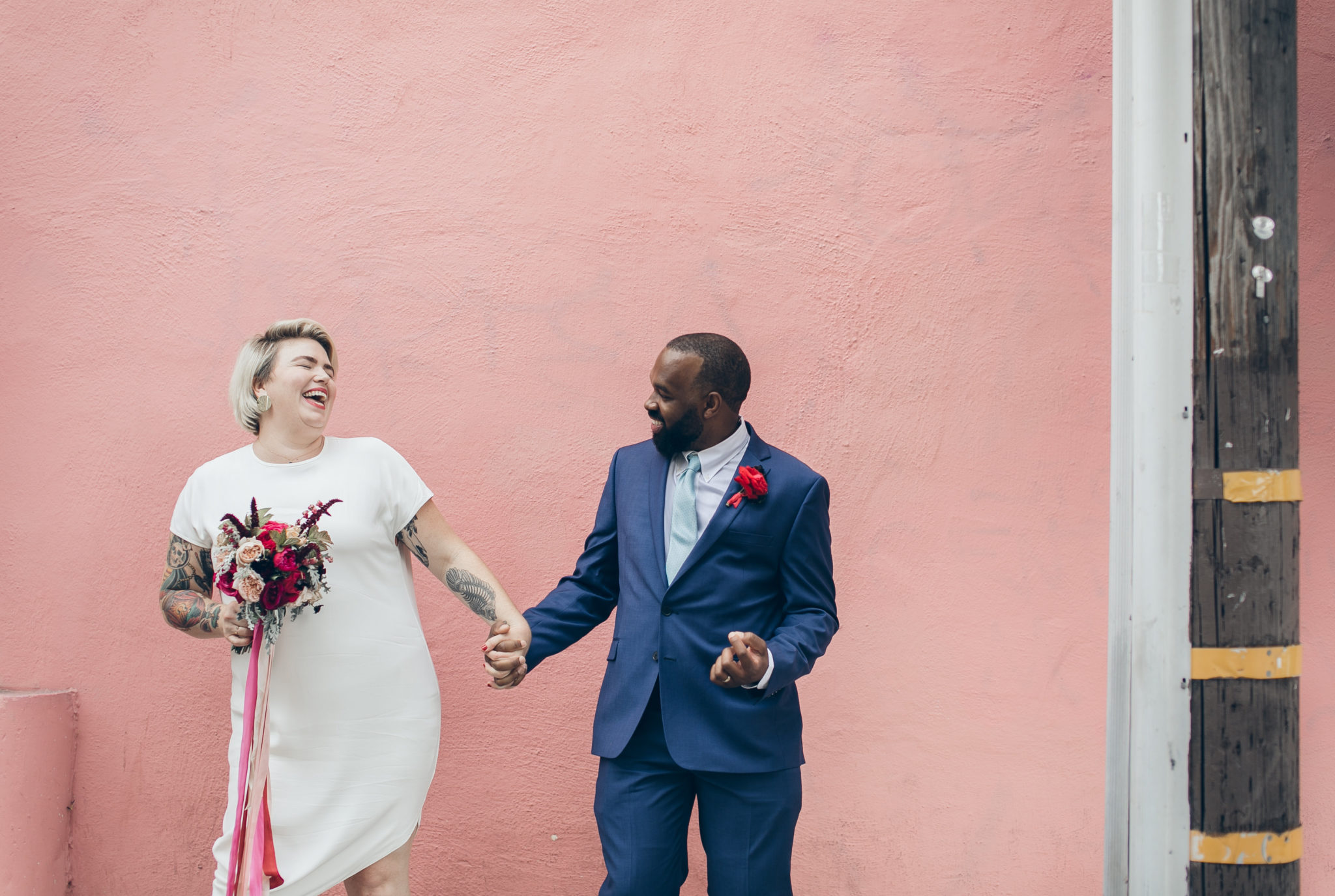 a plus size bride wearing a short wedding dress holding hands with a black groom in front of a pink wall