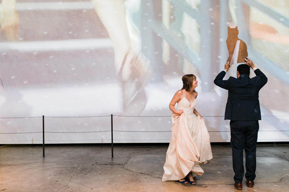 bride and groom dance in front of a wall with a large video projection while groom holds a larger than life cardboard cutout of a cat above his head in a photo by Laura Ford