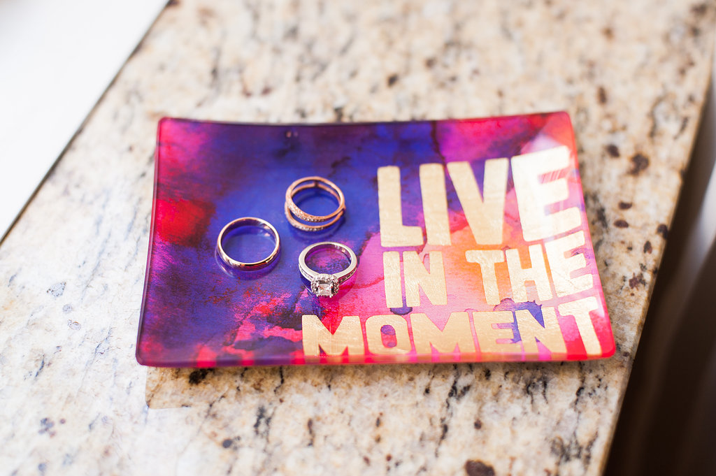 A small pink and purple jewelry dish reads "Live in the moment" in gold block lettering, as wedding bands and an engagement ring sit on the plate.