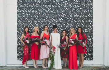 A bride in sunglasses and a cape dress stands among her six bridesmaids in different styles and shades of red dresses in front of a black and white number wall.