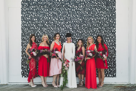 A bride in sunglasses and a cape dress stands among her six bridesmaids in different styles and shades of red dresses in front of a black and white number wall.