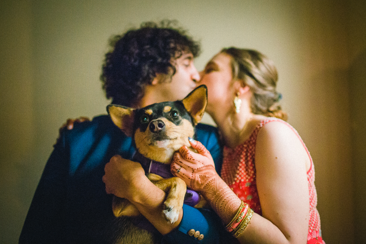 A couple kisses indoors on their wedding day while holding a small black and tan dog that looks away