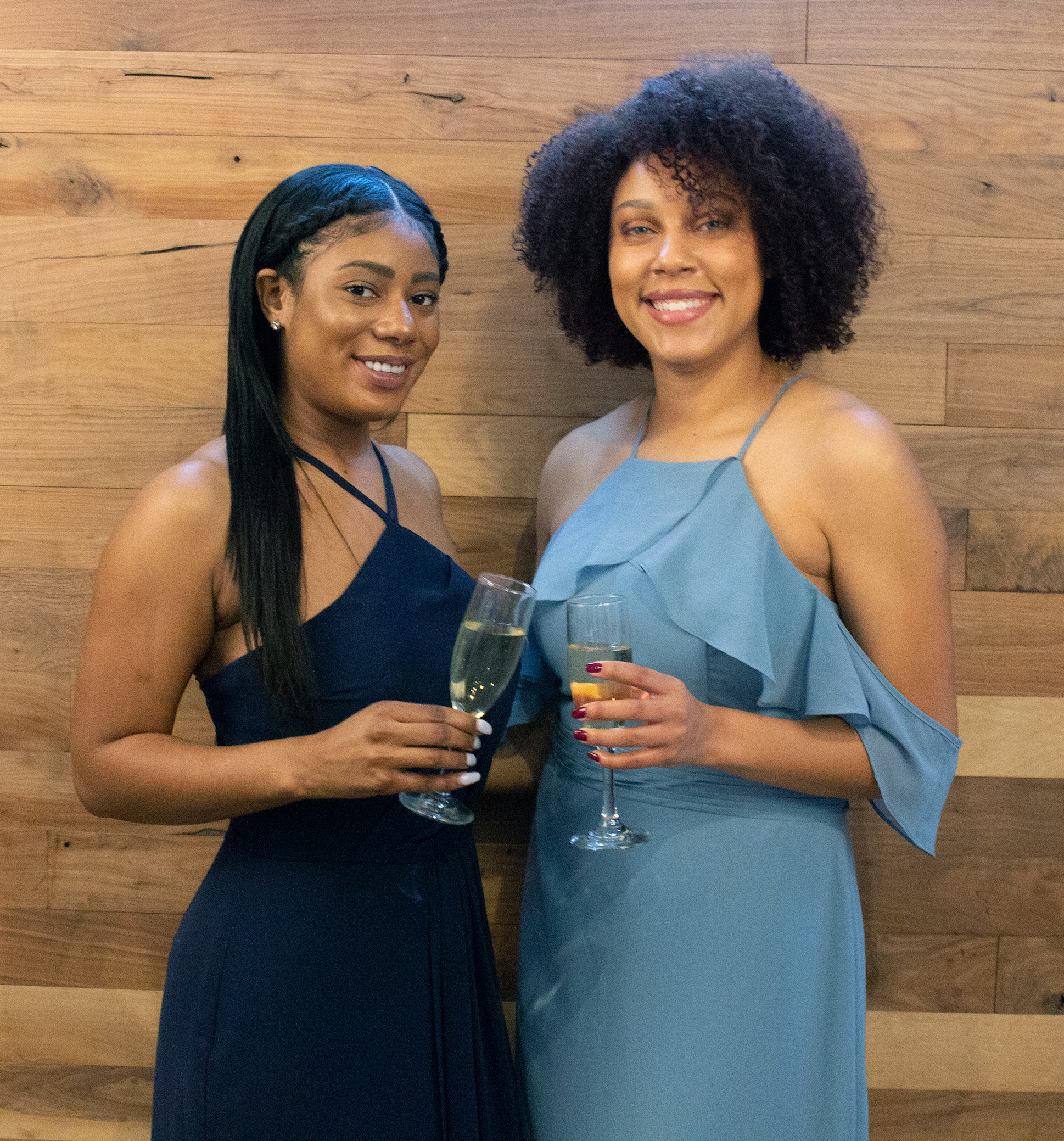 two women in blue Brideside bridesmaid dresses toast with champagne flutes in front of a wood wall