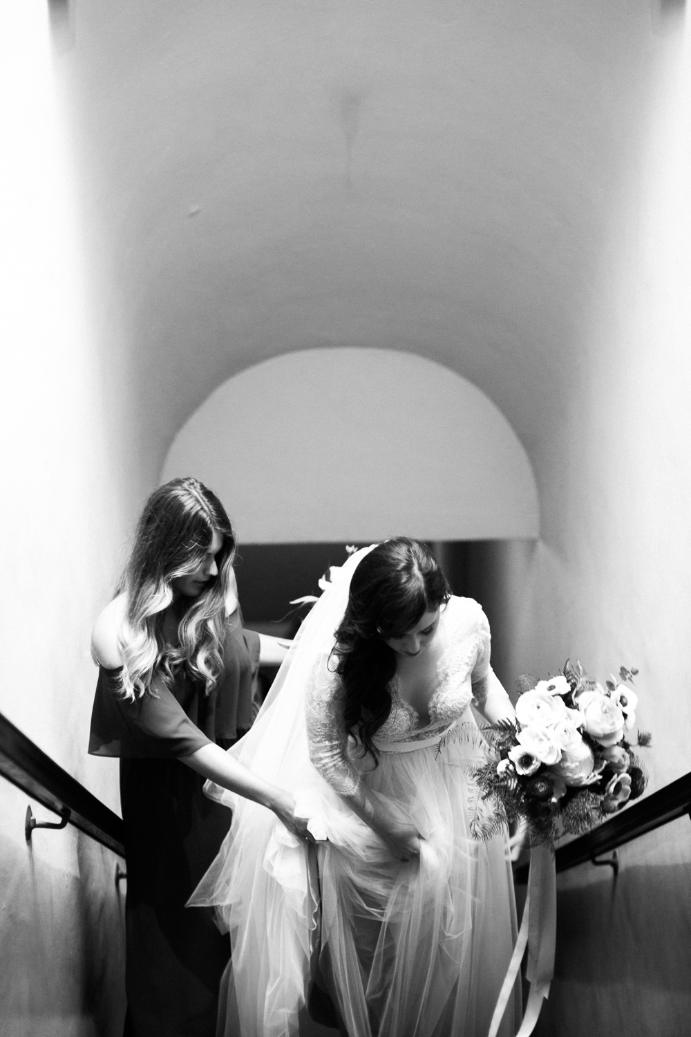 black and white photo of a bridesmaid helping a bride up a staircase framed by a barrel vaulted ceiling in a photo by Laura Ford