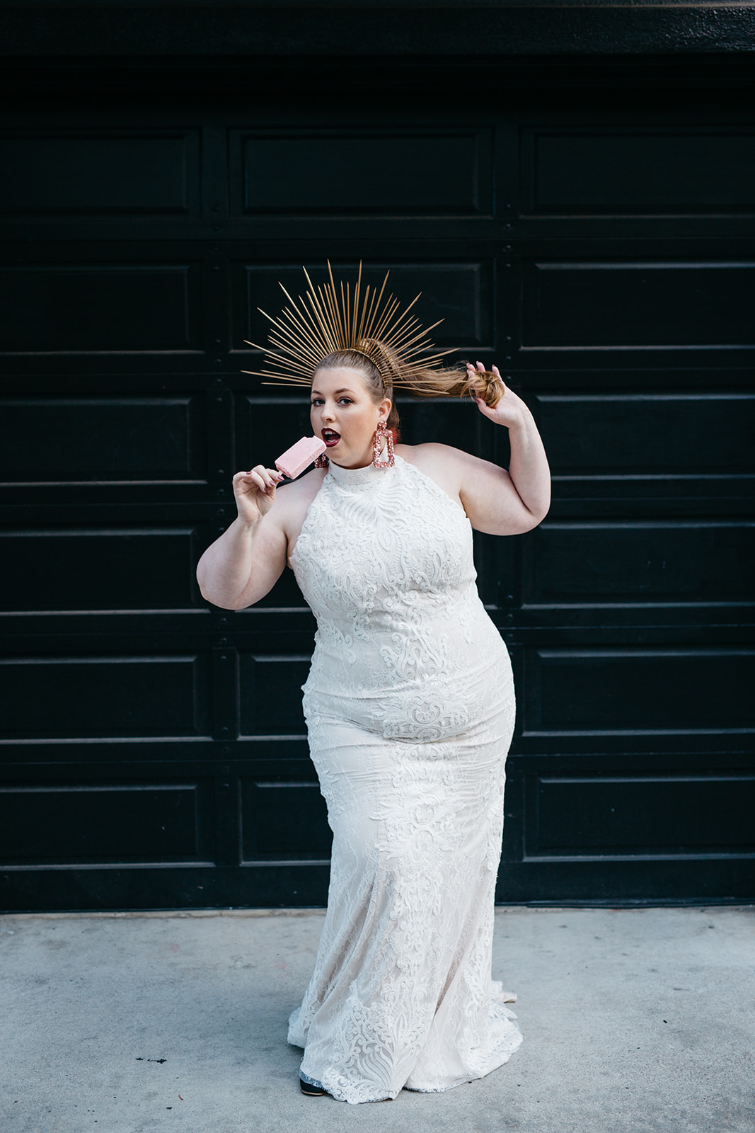plus size model wearing a high neck halter all over lace gown from the a practical wedding plus size wedding dress collection in front of a black wall and eating ice cream
