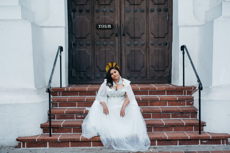 A woman in a radiant crown and a plus size wedding dress with a cape from Lace and Liberty x APW sits on terra cotta steps in front of brown wood doors