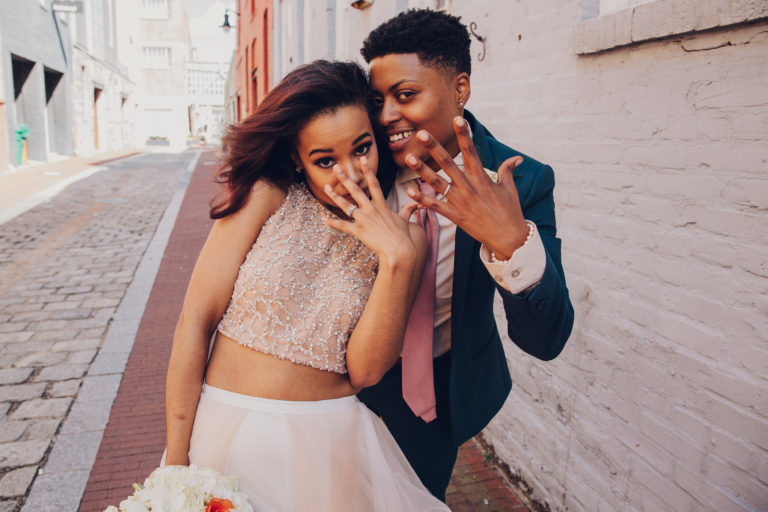 a queer couple show off wedding rings wearing a crop top wedding dress and a navy suit with a pink tie