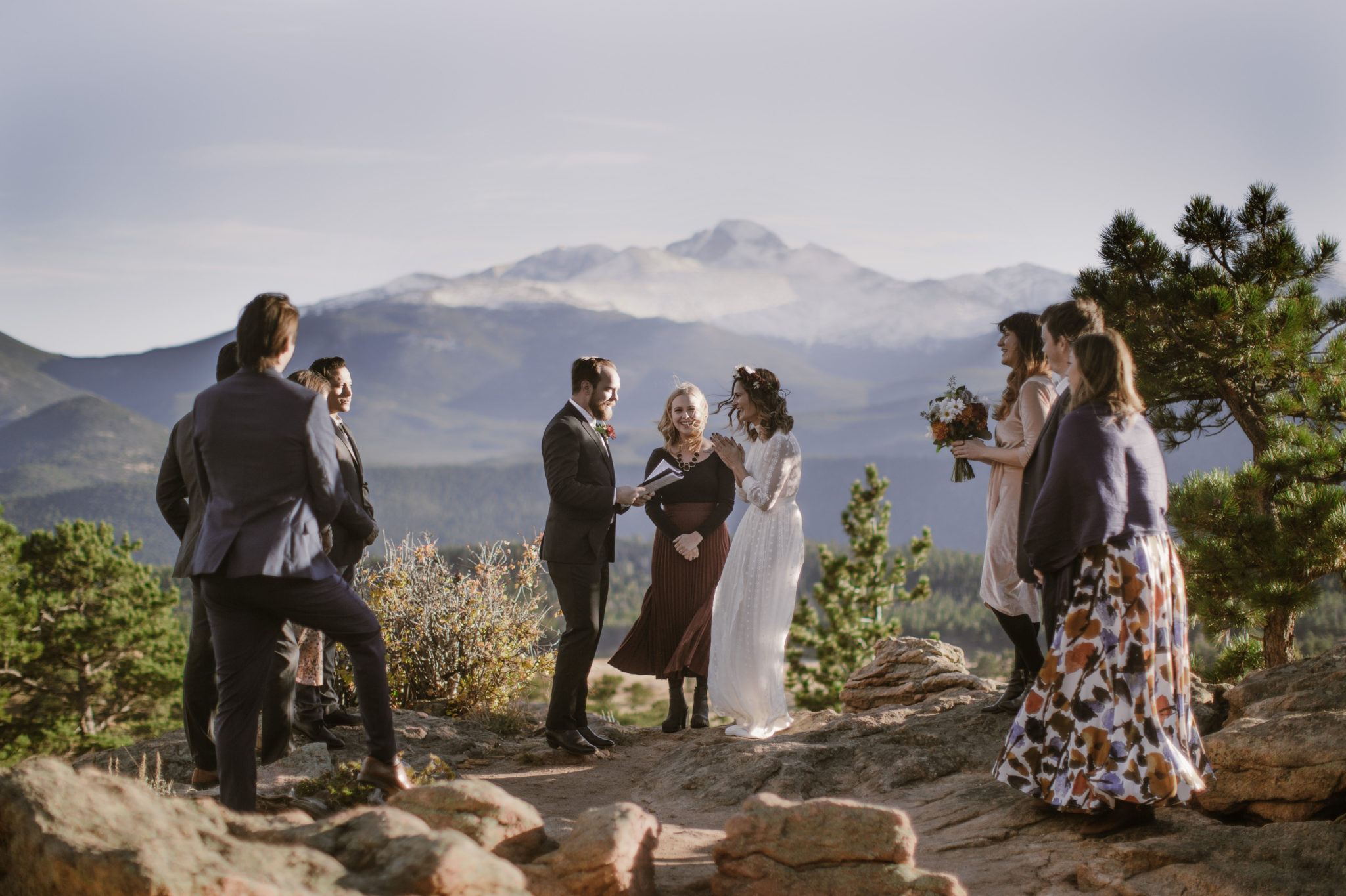 A couple in wedding clothes stands together near an officiant, being married on a mountain surrounded by a small group of people. Rocky Mountains form the backdrop in the distance.