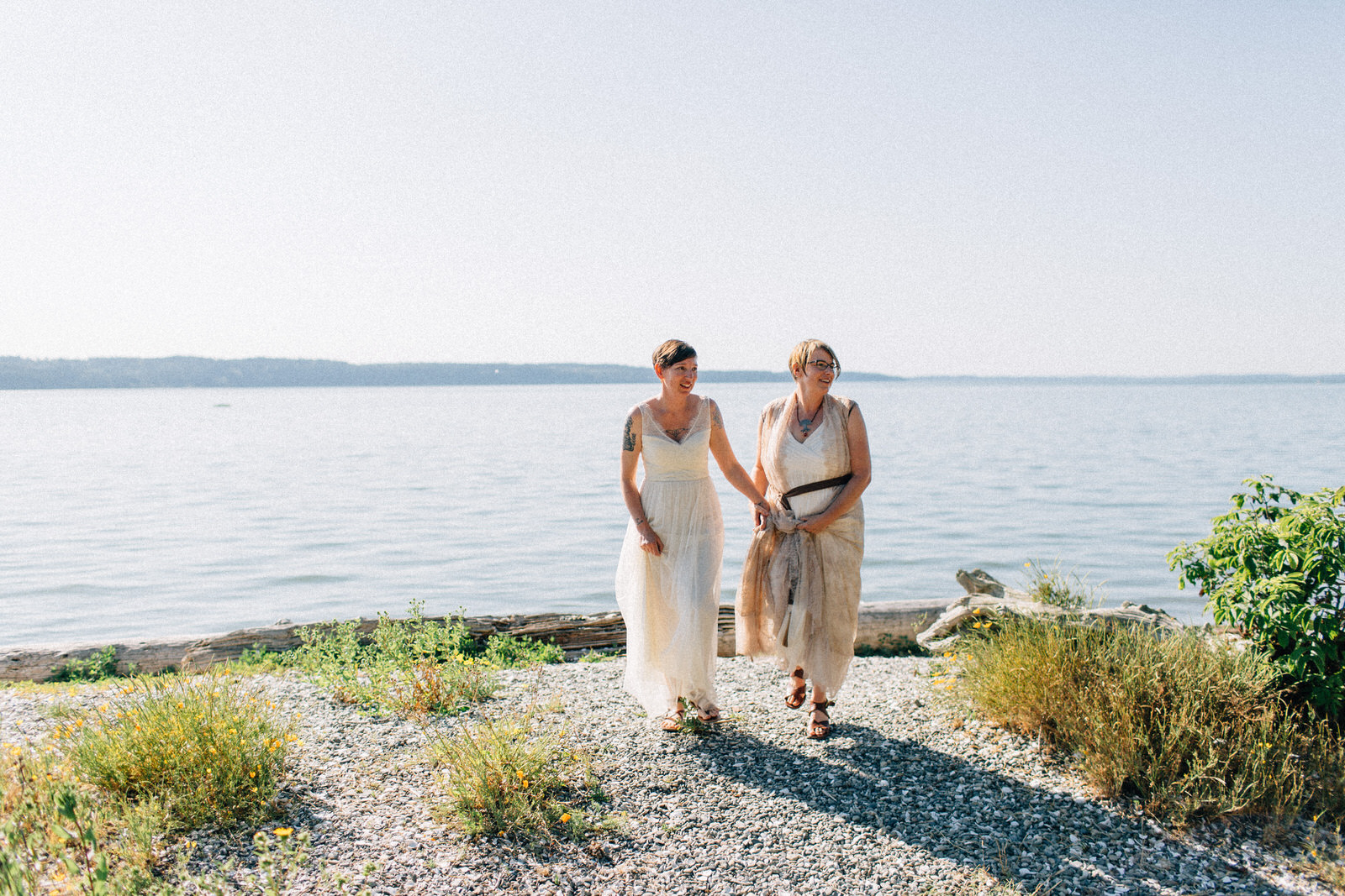 Two women in wedding dresses hold hands and walk along a pebbled beach, smiling