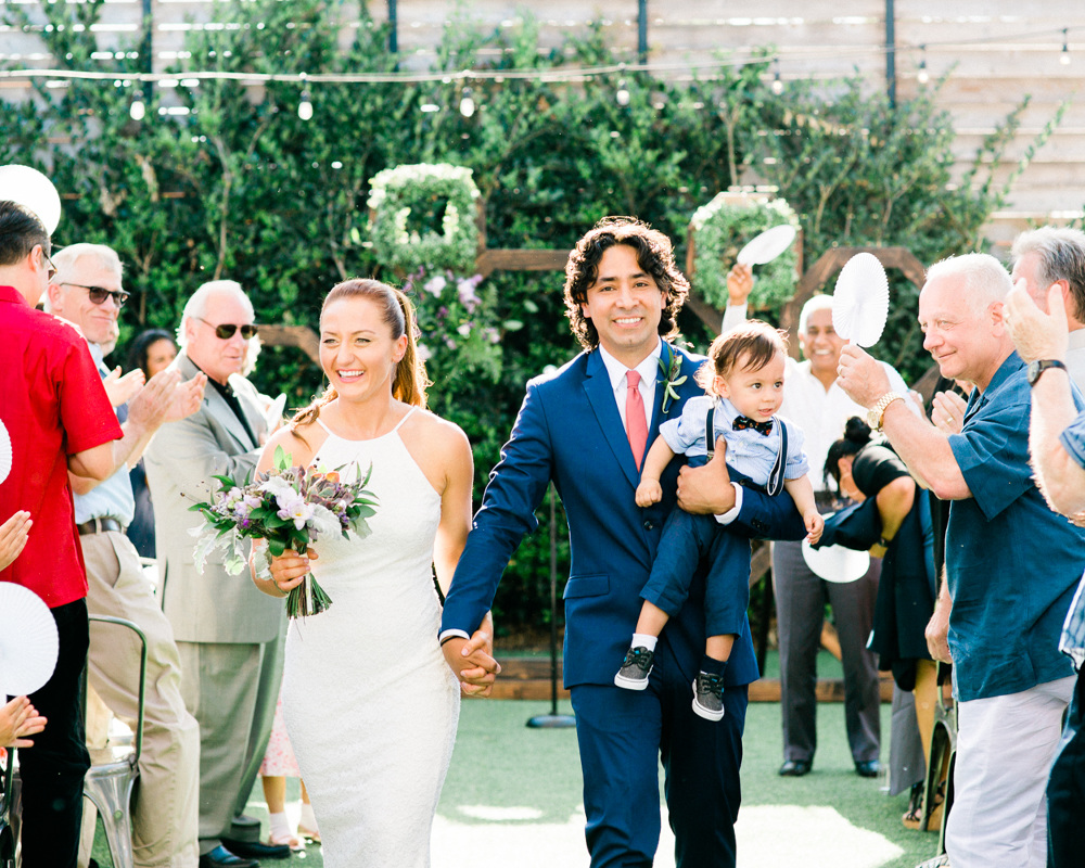 bride carrying bouquet and groom carrying small boy while walking down the aisle in a photo by Laura Ford